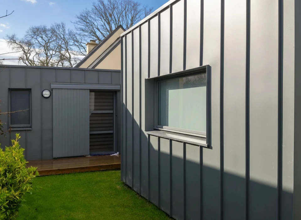 Extérieur résidentiel avec toiture en tôle ondulée métallique grise, cheminée en brique rouge, pignon à bardage vinyle horizontal bleu-gris avec ventilation circulaire blanche